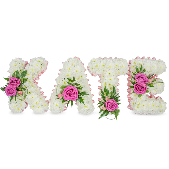 This deeply personal tribute reads as a floral portrait of the name KATE, fashioned in pronounced three-dimensional letters where every surface is a field of small, immaculate white chrysanthemums. The chrysanthemums are tightly packed to create a velvet-like, cloud-soft texture across each letter, while a delicate pale-pink satin ribbon traces the inner shapes, offering a subtle blush contrast that is both elegant and softly feminine. Strategic clusters of hot-pink roses break the white expanse with confident colour: two opened roses nestle along the left arm of the K, a single striking rose sits within the lower arc of the A surrounded by tiny pink buds and sprigs of green, the T is serenely anchored by one vivid rose at its base framed with fern-like fronds, and the E is brightened by two roses along its right face with variegated leaves and delicate pink accents threaded between. The greenery - ruscus tones, slender fern textures and glossy variegated leaves - gives the arrangement a garden-fresh vitality while preserving the dignity of the piece. Set against a pristine white background, the letters cast soft shadows that emphasise their freestanding structure and sculptural quality. The overall impression is luminous and restrained: tactile petals, a faint rose scent, and an intimate, sympathetic mood that suits a funeral chapel or celebration-of-life gathering. Local families across Grove Park, near Chinbrook Meadows and the quieter streets around Lewisham, often choose this style when they want a distinctly personal, readable tribute that balances tenderness with graceful craftsmanship.