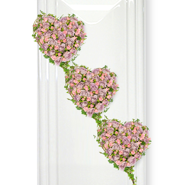 Three heart-shaped floral designs cascade gently down the frame in this sophisticated funeral tribute, each heart brimming with roses and edged with fresh foliage. The hearts are arranged diagonally, beginning near the upper left and descending gracefully to the lower right, creating a flowing visual path that feels like a gentle progression through cherished memories. Within each heart, small to medium-sized roses are tucked closely together, their petals overlapping in lush layers of dusty lavender, soft mauve, and pale blush pink. The colours blend in a harmoniously muted palette, evoking both romance and quiet reverence, reminiscent of the calm atmosphere along the leafy streets and riverside walks around Grove Park. Some roses appear newly opened, with crisp, cupped petals, while others are more mature, their layered forms fully unfurled, adding texture and a sense of time passing. Encircling and linking the hearts, delicate trails of ivy curl and twist, their slender stems and glossy green leaves forming a continuous outline and natural bridge between each floral cluster. This greenery provides contrast to the pastel tones, and also suggests the enduring ties between loved ones, even in bereavement. The background is bright, clean white, with faint vertical reflections that hint at glass or polished surfaces, like those you might see in a modern chapel or funeral home serving the Grove Park community. The lighting is soft and even, casting no harsh shadows and allowing every petal and leaf to stand out with clarity. The overall impression is one of elegant simplicity and deep emotion, perfectly suited for a tribute at a memorial service, cremation, or graveside farewell where family and friends want a symbol that speaks gently of lasting love and connection.