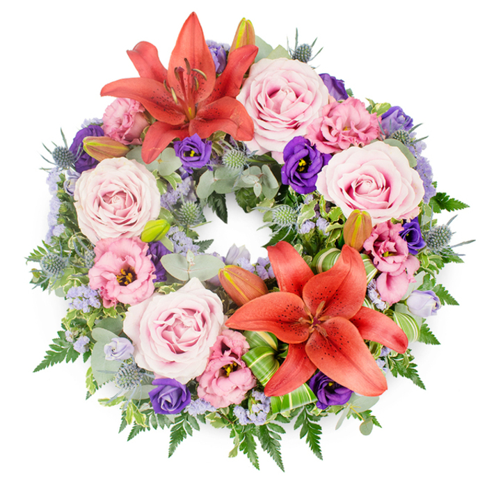 This image shows a meticulously arranged funeral wreath laid flat on a pure white surface, the neutral backdrop allowing its colours and craftsmanship to stand out with clarity. The wreath is perfectly circular and generously filled, built upon a lush base of varied greenery. Rounded eucalyptus leaves, graceful fern fronds, and deeper green foliage are interlaced to create a rich, layered framework that evokes the tranquil foliage-lined avenues of Grove Park. Resting upon this green collar, clusters of large blush-pink roses are positioned throughout, their soft, velvety petals spiralling open to full bloom and expressing a sense of affection and remembrance. Interspersed among the roses, lisianthus blossoms unfurl in soft pink and luxurious deep purple, each ruffled bloom adding contour, volume, and a gentle contrast of light and shadow. Two standout Asiatic lilies in a vivid reddish-orange tone act as focal points: one installed near the top of the wreath, the other placed toward the lower curve. Their wide, star-shaped petals and protruding stamens add structure and a touch of bold colour, symbolising courage and devotion. Scattered between these focal blooms are spiky blue eryngium thistles, lending a cool, textural counterpoint, and clusters of tiny lavender-blue filler flowers that cast a soft, mist-like veil across the arrangement. A few smooth, green lily buds, still closed, are tucked discreetly within the foliage, hinting at life continuing beyond the moment of farewell. Decorative, striped green leaves are artistically folded around the lilies, demonstrating precise, contemporary floristry. Overall, the wreath radiates calm, respect, and understated elegance, making it a thoughtful choice for funeral and memorial services in Grove Park, where Grove Park Florist can deliver such tributes to local churches, crematoria, or family homes.