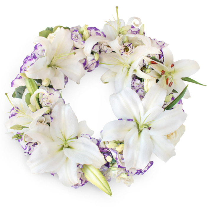 A dignified, circular funeral wreath is presented against a clean white studio background, its tranquil palette and careful structure creating an immediate sense of peace. The wreath is densely composed, with large white Oriental lilies forming the dominant visual theme. Their broad, satiny petals curve outward in open, star-like shapes, while a few blooms remain in partial bud, adding variation in size and shape. At the centre of each open lily, slender filaments rise up, bearing cinnamon-brown anthers that offer a gentle, earthy contrast to the cool whiteness of the petals. Threaded among the lilies are clusters of Lisianthus, their delicate, paper-thin petals twisting softly into ruffled cups. These blossoms appear mainly white, yet are brushed at the edges with lilac, lavender, and soft violet, adding a whisper of colour similar to the muted wildflowers found in the quieter corners of Grove Park and nearby Gunnersbury. Beneath and between the flowers, touches of deep green foliage and bluish, eucalyptus-like leaves peek out, giving the wreath a structured, slightly layered look without disrupting its airy feel. Tiny creamy buds are tucked in close to the lilies, lending an additional sense of fullness and continuity. The unbroken ring of flowers symbolises eternity and the enduring nature of memory, making this style particularly fitting for families arranging funeral tributes, memorial deliveries, or graveside flowers around Grove Park and West London. In the soft, even lighting, every petal texture is visible, from the smooth lilies to the more finely pleated Lisianthus, resulting in a wreath that feels both tender and composed, like a quiet moment of reflection before a service.
