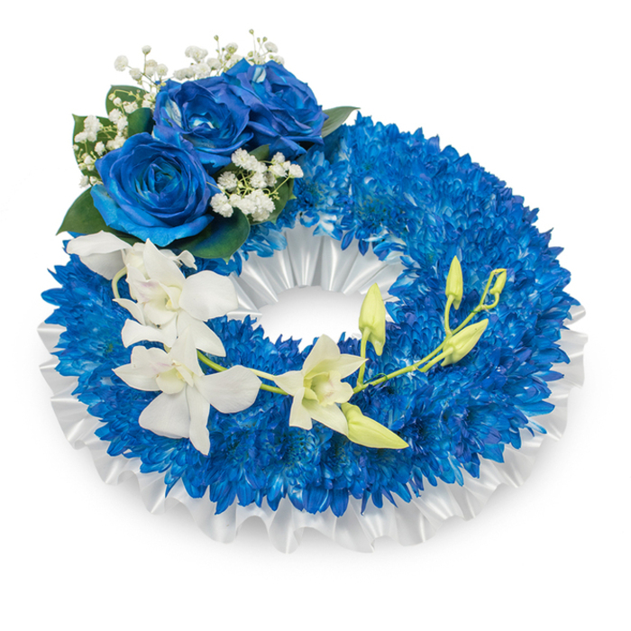 A refined circular funeral wreath in a thoughtful palette of deep blue and soft white, presented on a plain white surface to emphasise form and colour. The principal mass consists of innumerable small, vivid blue chrysanthemums tightly arranged to form a luxuriant, textured ring that feels dense and protective. Underneath and along the inner aperture a pleated white satin ribbon folds in gentle ruffles, its satin sheen creating a muted contrast that frames the floral ring like a quiet collar. Dominating the upper-left quadrant is an artful trio of large blue roses, petals unfurled in a velveteen sapphire hue that harmonises with the chrysanthemum base; around them, delicate clusters of baby's breath and touches of dark green foliage soften the transition between the dramatic blue and the cleaner white elements. Flowing from mid-left toward the lower-right is an elegant cascade of ivory-white orchids, their broad, graceful petals and subtle curves lending an ethereal, almost floating quality to the composition. Mixed among these orchids are lime-green buds-some closed, some just beginning to open-suggesting renewal even within a commemorative context. The overall composition balances stately structure with delicate movement, evoking serenity, respect and lasting memory. Imagined for services or graveside tributes in and around Grove Park-near Grove Park station and the local nature reserve-this wreath speaks of purity and condolence, crafted with the calm attention to proportion and finish expected from seasoned florists.