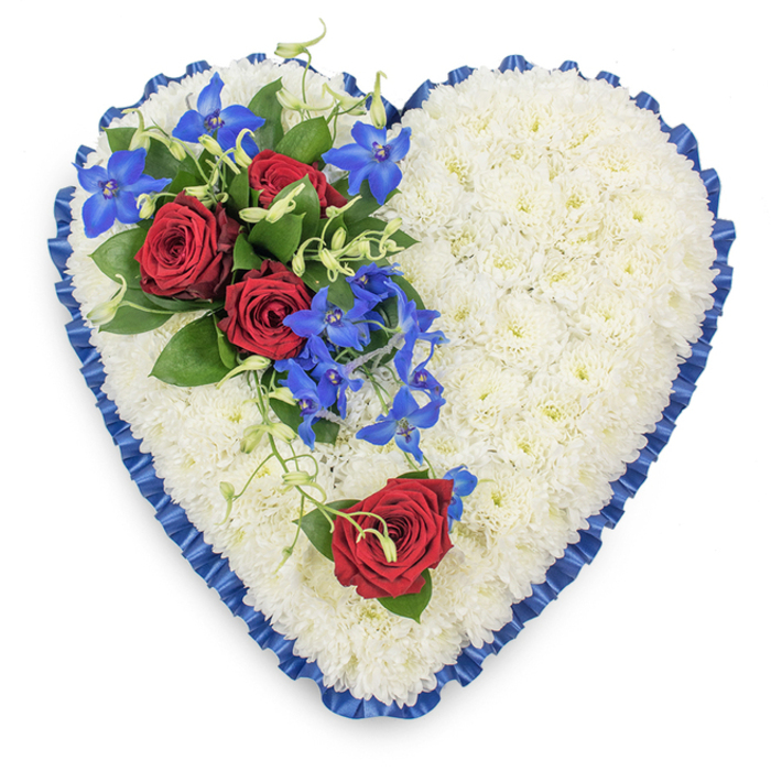 In this detailed top-down photograph, a large heart-shaped floral arrangement fills the frame, standing out clearly against a simple white background. The heart's entire surface is made from tightly massed white chrysanthemums, each small, daisy-like floret nestled next to the other to create a smooth, cushiony plane of soft texture. Their immaculate white petals lend the piece a serene, almost luminous quality. Around the perimeter, a royal blue satin ribbon has been carefully pleated and slightly ruffled, following every curve with precision. Its rich blue tone and subtle sheen define the heart's shape and bring an air of ceremony, similar to the calm, composed atmosphere of memorials held around Grove Park. On the upper left curve of the heart, a dramatic floral spray sits atop the chrysanthemum base. Three fully open deep red roses form the focal point, their velvety petals unfolding in layered spirals, framed by dark glossy foliage. Interlaced among them, electric blue dendrobium orchids introduce a striking contrast; their elongated stems and star-shaped blooms create vertical movement, as though the flowers are gently cascading down into the centre of the heart. Delicate green tendrils curl across the surface, softening the structure and adding a touch of natural whimsy. On the lower right arc of the heart, a single red rose is placed alone, mirroring the main cluster yet standing quietly apart, like one treasured memory held close. The overall palette of white, red, and blue speaks of purity, devotion, and peace, making this heart an eloquent tribute for a funeral or wake. It feels perfectly suited to a thoughtful farewell in the Grove Park area, where its calm beauty would offer comfort to grieving family and friends.