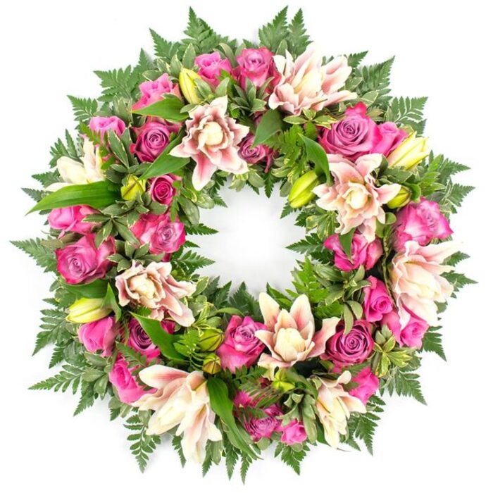 On a crisp white background, a beautifully composed circular wreath draws the eye with its rich mix of colour and texture, clearly intended as a funeral or memorial tribute. The wreath forms a complete, symmetrical ring, entirely filled with flowers and foliage so that the circle feels continuous and whole. At its heart are numerous roses in a spectrum of pinks, from intense fuchsia and magenta through to softer, romantic blush tones. Some of the roses remain in tight buds, their petals carefully wrapped, while others are half-open or fully open, showcasing layer upon layer of soft, velvety petals that seem to fold and curl inwards. Nestled between these are lush double lilies, their petals arranged in multiple layers and opening outwards in hues of creamy white, pale blush, and light peach. Subtle touches of darker pink along the petal edges add a gentle sense of depth. Scattered among the open blooms, lime-green lily buds stand upright, their pointed tips offering a fresh counterpoint to the surrounding softness. All of this is supported by an abundant base of greenery: fine fern fronds create an airy, feathered border around the wreath, while glossier, broader leaves sit close to the flowers, their deep green tones providing a rich contrast that allows the roses and lilies to really stand out. The white setting gives the impression of the wreath resting on a florist's bench in Grove Park, freshly assembled before being taken to a service or laid at a graveside. The overall mood is one of gentle elegance and respectful remembrance, an arrangement designed to bring a quiet, soothing beauty to moments of farewell.