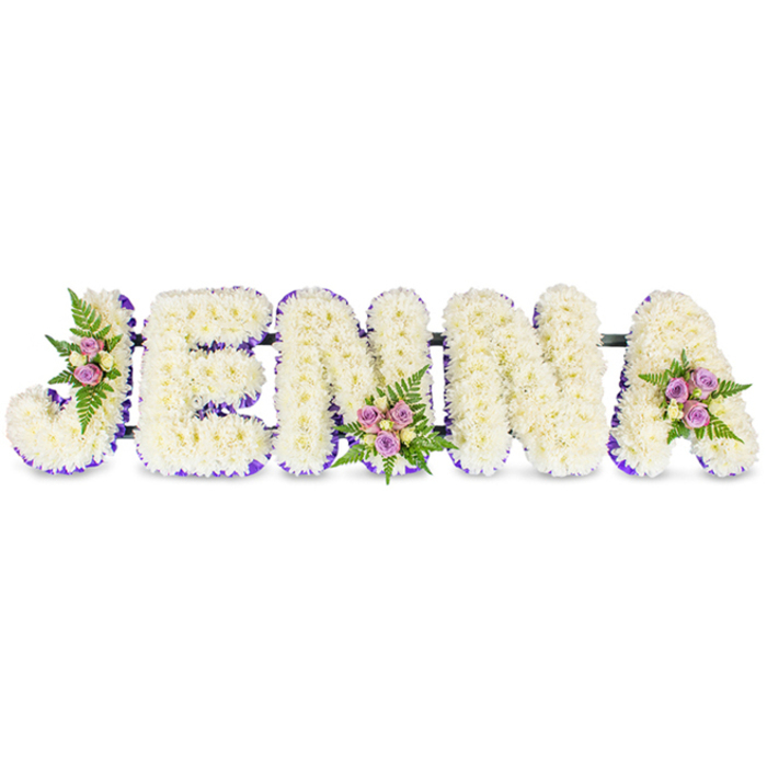 A lovingly assembled memorial commission spells the name JENNA in large, three-dimensional capitals fashioned from hundreds of tiny, creamy white chrysanthemums. Each letter feels almost cloudlike to the eye, with dense, rounded heads creating a comforting, tactile surface that softly reflects ambient light. A rich purple trim - appearing as fine ribbon or closely set lavender blooms - outlines the internal contours of each letter, supplying a measured contrast that sharpens the typography without overpowering the serene whites. Three small, artfully placed floral clusters enliven the layout: one on the upper left curve of the J, another at the lower middle of the first N, and a third on the upper right sweep of the A. Those clusters pair delicate blush and lavender spray roses with pale cream buds and sprigs of deep-green fern, introducing subtle colour, a hint of rose fragrance, and layered texture amidst the chrysanthemums. Slim, dark metal bars discretely connect the letters, evidencing the careful mechanical support required for a stable tribute. Intended as a sympathy or funeral arrangement, this piece is ideal for a chapel display, service table, or graveside offering - crafted with the steady hand and quiet attention to detail familiar to florists working in Grove Park. At Grove Park Florist we prepare such bespoke name tributes to be easily placed at wakes or memorials, and we consider local delivery to nearby streets such as Baring Road and areas around Grove Park station when arranging service logistics.