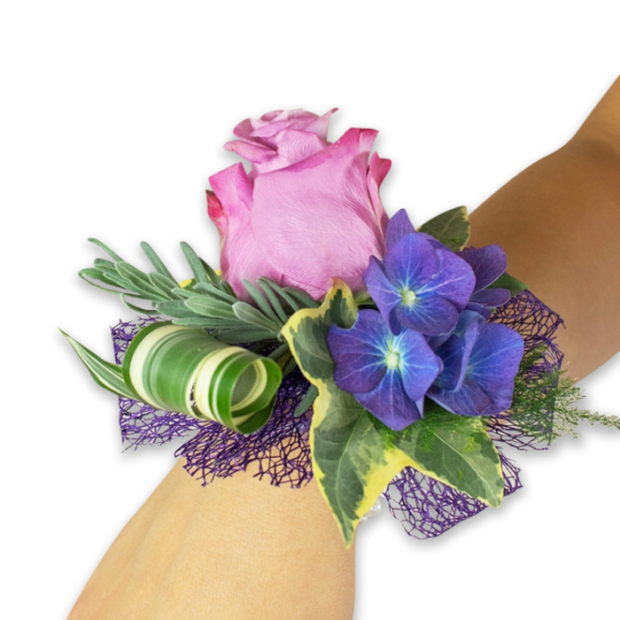 A meticulously crafted wrist corsage sits on a pale forearm, photographed against pristine white to emphasise its colours and textures-an understated jewel from a neighbourhood florist near Grove Park station. Dominating the design is a generous, partially open pink rose whose petals graduate from a warm fuchsia at the ruffled outer edge to a softer blush toward the heart, creating a romantic focal point that feels both classic and intimate. Flanking the rose are three rich clusters of deep indigo hydrangea; each tiny floret carries a luminous bluish centre that adds dimensional sparkle and a velvety depth to the arrangement. Glossy variegated ivy leaves provide a timeless border, their dark green centres edged with creamy highlights, while delicate silver-green sprigs-evocative of eucalyptus-fan behind the blooms to add a soft, aromatic backdrop. A statement leaf with dramatic white and green striping has been curled into an elegant spiral, introducing modern, architectural movement that contrasts beautifully with the natural softness of the rose. A layer of dark purple open-weave mesh beneath the flowers peeks through, giving texture and shadow that amplifies the hydrangeas' tone. The piece balances silky, crisp, and slightly coarse textures, and suggests a faint floral-herbal scent under warm, diffused light. Crafted for special occasions-proms, weddings, or an anniversary night out-this corsage is the sort of thoughtful, locally made detail proudly arranged by Grove Park Florist for clients across Grove Park, Hither Green and Lee.