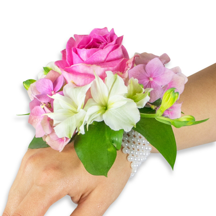A refined floral wristlet is photographed on a left hand and forearm with a light-to-medium skin tone, posed against a spotless white surface to highlight every tactile detail. The focal point is a large, fully opened deep fuchsia-pink rose, its layered petals dense and slightly cupped, offering a plush, velour-like texture at the heart of the arrangement. Surrounding the rose, clusters of blush-pink hydrangea florets lend a soft, voluminous backdrop, their tiny petals overlapping like silk to create a gentle halo. Scattered throughout are crisp white alstroemeria (Peruvian lilies), each with four to six delicate petals and faint light-green streaks near the throat, bringing clarity and a bright counterpoint to the richer pinks. On the right-hand side a grouping of pale green alstroemeria buds and a few almost-closed pink buds introduce visual movement and the suggestion of a blossom about to open. Broad, glossy emerald-green leaves cradle and lift the composition, their smooth, reflective surfaces framing the flowers and enhancing the colour contrast. The ensemble is secured to the wrist by an elegant, multi-strand bracelet of small, luminous white pearls that wraps neatly around and extends partially down the back of the hand, adding a touch of heirloom grace. This corsage reads as an emblem of intimate celebrations-wedding parties along local Grove Park streets, graduation ceremonies, or prom evenings-combining scent, texture, and expert assembly with a calm, wearable sophistication.