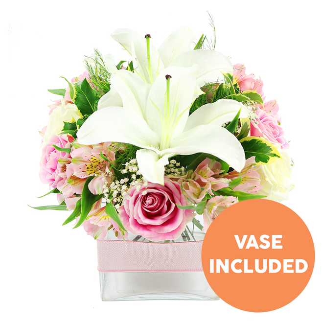 This image presents a meticulously arranged floral display against a pure white backdrop, the kind of bouquet that would feel perfectly at home on a windowsill overlooking Grove Park's tree-lined streets. In the centre, two large Oriental lilies in pristine white command attention, their open petals arching outward to reveal textured, reddish-brown stamens that rise from the heart of each bloom. These lilies are surrounded by a generous halo of pink roses, some just beginning to unfurl, others already fully opened so that their petals form dense, layered rosettes. The pink tones are soft and romantic, shifting gently between blush and a slightly deeper hue, evoking warmth and affection. Threading through this central mass are slender stems of alstroemeria in complementary shades of pink and white, their elongated petals delicately flecked with yellow and fine speckling, adding a sense of movement and fine detail that rewards close inspection. Scattered among the larger blooms, tiny clusters of white baby's breath create a light, gauzy texture, almost like a veil of mist softening the whole arrangement. A mix of lush green foliage forms the backdrop and frame: broad, glossy leaves provide structure and contrast, while finer, feathery greenery suggests a more natural, garden-picked feel, reminiscent of the plant life around nearby Chiswick House and Gardens. All of the flowers and leaves are gathered neatly into a clear, square glass vase, its simple lines allowing the stems and clear water to be seen, reinforcing the impression of freshness. Around the centre of the vase, a pale pink satin ribbon is tied in a smooth band, lending a gentle, gift-ready polish. In the bottom right corner, a bright orange circular badge stands out on the white background, with bold white capital letters stating VASE INCLUDED, making the presence of the vase unmistakably clear. The bouquet as a whole conveys elegance and tenderness, suited to occasions from anniversaries and birthdays to simple gestures of appreciation in Grove Park.