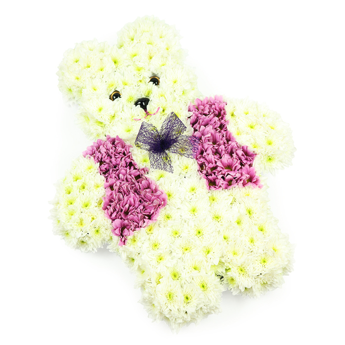 This photograph showcases a lovingly designed teddy bear floral tribute crafted by a skilled florist familiar with the quiet, leafy character of Grove Park. The bear is seated upright against a completely white background, its whole shape formed from creamy white chrysanthemums packed so closely that barely a stem can be seen. The petals overlap to create a thick, textured coat that looks as though it would be soft beneath the fingertips, bringing to mind a favourite teddy resting on a bed in a house near Fauconberg Road. The head is smoothly rounded, with distinct ears that curve gently outwards, framing a face that feels disarmingly gentle. Two small, dark shapes mark the eyes, and a slightly larger dark element forms the nose, setting off the pale petals and giving the bear an expression of quiet, steadfast kindness. The arms curve slightly inwards, as if the bear is ready to offer a hug, while the legs are short and sturdy, stabilising the form. Draped across the chest is a floral waistcoat made from deeper pink and mauve flowers that introduce richness and depth to the design. These blooms are more open and voluminous, creating two neat panels that meet at the centre. Just where the vest meets the neck, a bow of sheer purple netting sits proudly, its gauzy folds adding lightness and a touch of ceremony. The lighting in the image is bright yet soft, allowing the white chrysanthemums to glow gently without glare, and ensuring that the subtle interplay of colour and texture is easy to discern. Intended as a funeral or memorial tribute, especially meaningful for a child or someone with a fondness for soft toys, this floral teddy would bring a sense of tender remembrance to services across Grove Park, Chiswick, and the surrounding riverside neighbourhoods.