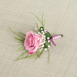 A delicately crafted boutonniere is displayed against a natural, light beige linen backdrop, the subtle weave of the fabric giving a relaxed, homespun charm that feels typical of intimate Grove Park weddings and proms. At its heart sits a single blush pink rose, captured in that lovely moment between bud and full bloom. The centre is a gentle, powder-soft pink, while the petals deepen to a more vibrant rosy shade as they curl outward, each velvety layer catching the light and hinting at a soft, classic fragrance. Surrounding the rose, clusters of creamy white baby's breath form a light, frothy collar, their tiny blossoms adding texture and a romantic, almost vintage feel that many local couples request when visiting Grove Park Florist. Behind the central bloom, slim, grass-like strands of fresh green foliage extend in gentle arcs, lending movement and a sense of airiness, like wisps of garden greenery gathered from a nearby park. A single, broader dark green leaf is folded neatly around the base, creating a clean, tailored shape that keeps the small arrangement tidy and lapel-ready. The stems are tightly bound in a smooth satin ribbon, a blend of soft lavender-pink and richer magenta-purple tones that shimmer subtly under the light, tied into a simple, secure knot. The colour of the ribbon harmonises with the rose while adding a hint of contemporary style suited to modern ceremonies across Grove Park and surrounding areas. Soft, even lighting highlights the contours of each petal, the delicacy of the baby's breath, and the understated elegance of the linen, turning this modest boutonniere into a refined statement piece for weddings, proms, or formal celebrations.