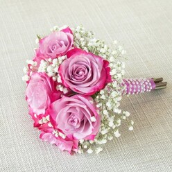 In the heart of Grove Park, a classically inspired wedding bouquet sits poised on a soft, neutral linen, capturing the fresh, joyous spirit of local celebrations. Five radiant roses form the bouquet's centerpiece; their petals reveal a mesmerizing two-tone transition, dusk lavender blossoming into bold fuchsia at the outermost edges. The composition gains a whisper of airiness from countless white baby's breath florets, which seem to float beside the opulent roses. The expertly wrapped stem-deep plum satin, meticulously wound with pearlescent white beads-offers the tactile intrigue of elegance and refinement, mirroring celebratory mornings along Grove Park Road. Beneath the ribbon, freshly trimmed green stems peek through, suggesting a just-gathered charm. Bathed in gentle natural light, the bouquet's colours sing against the tranquil backdrop of subtle, oatmeal fabric. Crafted by Grove Park Florist for local weddings, anniversaries, or special occasions, this sophisticated yet welcoming bouquet carries the romance of riverside walks and heartfelt gatherings, making it perfect for sharing the beauty of Grove Park with those you love.