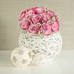 A beautifully styled still life captures a rounded bouquet of pink roses and white baby's breath, presented with the care of an experienced Grove Park florist arranging flowers for a special occasion. The roses, around fifteen in number, are tightly massed into a dome, each head at peak openness so that the subtle ombré of the petals is visible: pale, almost porcelain pink at the centre, warming gradually into a deeper rose and finally to a rich, bordering-on-fuchsia edge. They look plush and velvety, the kind of blooms that would feel cool and silky if you brushed them with your fingertips. Threaded through the roses are feathery clusters of white Gypsophila, the tiny blossoms hovering lightly between the larger blooms like mist, giving the entire arrangement an airy, romantic halo. The bouquet is nestled in a spherical white vase made from overlapping strips of material that mimic white-painted wicker or rattan, creating an open lattice pattern that lets the ambient light filter through. It sits on a pristine white surface that reflects the soft illumination, suggesting a bright windowsill or console table in a Grove Park townhouse. To the left, two decorative natural-fibre spheres in pale beige, one larger and one smaller, add a relaxed, organic touch, their open weave echoing the design of the vase. The background is a warm off-white wall, gently textured so it feels homely rather than stark. The whole composition radiates calm elegance and quiet celebration, perfect for marking an anniversary, welcoming a new baby home to Grove Park, or simply bringing a touch of refined softness to everyday interiors.