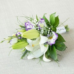 A refined, elongated bouquet rests at a gentle diagonal on a pale beige, woven fabric, its understated backdrop reminiscent of a calm Grove Park townhouse interior. The eye is first drawn to the white lilies that anchor the composition, their broad petals opened wide like starbursts, revealing prominent yellow stamens dusted with warm pollen and delicate freckles of russet along the inner throats. These lilies feel both serene and celebratory, as if poised to accompany a bride walking out of a local church or registry office nearby. Along the spine of the bouquet, several bright green lily buds stand upright, smooth and tapered, quietly suggesting the promise of blossoms still to come. Interlaced with the lilies are frilled Lisianthus blooms: their petals are soft and ruffled, white at the base and finished with vivid purple rims, some a gentle lilac, others a saturated violet. They bring a touch of modern colour to the otherwise classic palette. Sprigs of airy white baby's breath punctuate the design, creating a cloud-like softness that fills the spaces and adds a romantic haze. Surrounding it all is a lush foundation of greenery-broad, glossy deep green leaves that offer weight and contrast, intertwined with slender, more whimsical fronds and slightly spiky accents that give the bouquet an organic, freshly gathered-from-the-garden feel you might associate with local homes near Grove Park station. The gathered stems are tightly bound with a crisp white satin ribbon, discreetly embellished with tiny pearl-like beads glinting along one side, reinforcing the bouquet's suitability for weddings, anniversaries, or milestone celebrations. Soft, even light falls across the flowers, highlighting every texture from smooth petal to shining leaf, capturing a sense of freshness and care that reflects the quiet elegance of this London neighbourhood.