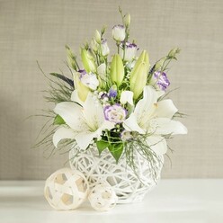A beautifully balanced floral display featuring white lilies and lisianthus is captured in gentle daylight, as if placed on a wedding table in one of Grove Park's characterful riverside venues. The arrangement is anchored by a distinctive round white vase whose open latticework resembles woven rattan, giving a sense of weightless structure while adding tactile interest. Emerging from this base, several tall white lilies command attention; some are fully open with wide, star-like petals and clearly defined stamens, while others remain in sleek, pale green buds that lend height and a sense of unfolding time. Clustered at mid-level, layers of lisianthus form soft, ruffled blooms in creamy white, many tipped with a delicate wash of lavender or lilac along their edges, like petals lightly brushed with watercolour. Interwoven among them are tiny white blossoms akin to gypsophila and a scattering of deeper purple or blue-toned florets that create gentle points of contrast. Lush greenery, including feathery fern-like fronds and slightly broader leaves, spills and arches over the rim, softening the outline and suggesting the easy, natural beauty of a well-tended Grove Park garden. On the polished white surface beside the vase, two decorative spherical ornaments made from light wicker or rattan rest side by side, echoing the open-weave texture and enhancing the cohesive, contemporary feel. Behind the arrangement, a light beige wall with a subtle horizontal, linen-like grain provides a calm, neutral backdrop, allowing the whites and greens to glow. The mood is one of quiet celebration and reassurance, making this design ideal for wedding receptions, baptisms, or heartfelt condolences, with local delivery available throughout Grove Park and neighbouring streets.