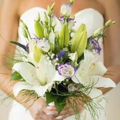 Grove Park's romantic energy is distilled in this luxurious bridal bouquet, held protectively by a bride on her special day. Exuding scent and grace are the mainstay Oriental lilies: their snowy, trumpet-shaped blooms wide open to catch the soft daylight, each petal like a stroke of silk. Nestled alongside, lisianthus unfurl in creamy white with ruffled edges touched by gentle lavender, balanced by tighter buds whose eventual opening promises the day's new beginnings. Vivid purple Veronica spikes rise confidently from the composition-a bold tribute to the vibrant flower displays common at home gardens near Sutton Court Road. Clouds of tiny white gypsophila give a dreamy, ethereal softness. Lush green foliage, including unopened lilies and elegant asparagus fern, drape artfully, providing organic structure that echoes Grove Park's famous tree-lined avenues. The bride's well-groomed hands, with pale manicure shining, hold the bouquet tightly, while her strapless dress offers a serene, white canvas in the background. Every detail speaks of local craftsmanship-an arrangement thoughtfully created for weddings across Grove Park and nearby Chiswick, promising fresh beginnings rooted in the community's floricultural tradition.
