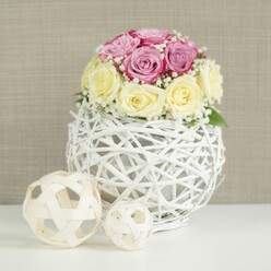 A delicately styled rose arrangement sits in the centre of the image, exuding the quiet elegance of a Grove Park townhouse prepared for an intimate celebration. The flowers are arranged in a compact, rounded bouquet, with approximately a dozen roses gathered closely together. Half of the blooms display soft shades of pastel and lavender-toned pink, while the others show creamy yellow and ivory tones, the colours blending gently like evening light by the River Thames. Each rose reveals velvety petals layered in tight spirals or gently unfolding whorls, conveying freshness and care in the way they have been selected and placed. Fine stems of white baby's breath thread through the roses, their tiny, cloudlike clusters adding a romantic haze and helping to fill every small gap without overwhelming the pastel palette. The bouquet is nestled into a memorable white vase shaped like a sphere, constructed from thin, branch-like strips of wicker or rattan painted crisp white. This open-weave design forms a delicate lattice or cage, allowing glimpses of the container within and giving the piece a sculptural, handcrafted feel. Resting on a smooth, white tabletop, the vase is accompanied to the left by two decorative woven orbs made from pale, natural wood. One orb is slightly larger and positioned just behind the smaller one, their airy, interlaced structures echoing the vase's texture and adding a sense of layered composition. In the background, a soft beige or taupe surface, perhaps a linen-like wall or subtle wallpaper, fades gently out of focus, ensuring no distraction from the roses and their unusual base. The overall effect is calm, inviting, and refined, like a table piece chosen from Grove Park Florist for a wedding breakfast, engagement party, or chic at-home gathering that calls for a touch of local floral artistry.