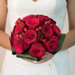 Under the soft glow familiar to Grove Park's garden venues, this bridal bouquet radiates a poetic blend of drama and sophistication. Every deep red rose in this arrangement appears handpicked, their textured petals creating a sense of movement and depth-some buds promising freshness, others unfolding with a classic grace. Scattered throughout are clusters of glistening red hypericum berries, whose rounded forms offer playful contrast, like punctuation in life's meaningful moments shared along Grove Park's bustling streets. Wrapped in a collar of broad, shiny green leaves, the bouquet is cradled with ease by the bride, whose white lace-trimmed gown shimmers in the gentle light of the occasion. Her neatly manicured hands gently support the bouquet, accentuating its significance and the company of close loved ones. The scene evokes a sensory blend of soft fragrance and tactile richness, conjuring images of heartfelt vows exchanged at intimate Grove Park ceremonies. This floral arrangement captures not only elegance and unity, but also the vibrant character of the local area-a signature piece for a day marked by love, joy, and the skills of Grove Park Florist.