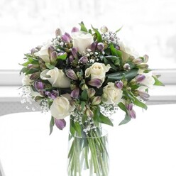 A delicately composed floral arrangement of white roses, lavender alstroemeria, and feathery baby's breath rests in a tall, cylindrical glass vase, bathed in soft, diffused daylight that suggests a bright window in a Grove Park home. The central roses are fully open, each bloom formed from layers of creamy white petals that curl outward with a smooth, almost satin-like texture, inviting the viewer to imagine their subtle, fresh fragrance. Clustered among them are plentiful stems of alstroemeria in muted shades of lilac and dusky purple, some still tightly closed, others just beginning to part their petals, showing fine streaks and veins of deeper colour that provide gentle contrast. Fine sprays of gypsophila scatter between these larger shapes like tiny white constellations, giving the bouquet a light, airy feeling and softening the transitions from one bloom to the next. Rich green foliage encircles and supports the flowers, from glossy, broad leaves at the base to more slender greens that weave upward, framing each head with a natural, garden-like ease. The long green stems are visible through the crystal-clear water in the vase, aligned with quiet precision and giving a sense of care and craftsmanship in the way the arrangement has been built. The overall impression is calm, elegant, and reassuring, reminiscent of a tranquil walk along the residential streets near Grove Park station or through the nearby river paths. It feels suited to sending thoughtful sympathy, celebrating an engagement, or simply bringing a sense of serenity and refinement into a living room, crafted with the attentive eye of an experienced local florist.