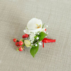 A single, elegant boutonniere is photographed from above against a subtly woven, oatmeal-coloured fabric, its gentle texture evoking a relaxed yet refined setting in Grove Park. At the centre sits a flawless white Phalaenopsis orchid, the petals broad, silky, and gracefully arched as if caught mid-breath. The orchid's throat glows with a wash of creamy yellow and muted orange, adding depth and warmth, like late afternoon light along the Thames. Surrounding the orchid, a halo of fine baby's breath blooms forms a gauzy, cloud-like ring of tiny white stars, each floret lending softness and an almost whisper-light texture. Tucked alongside are plump, glossy red berries that feel rich and festive, their rounded shapes offering a playful contrast to the orchid's smooth planes. A few fresh green berry buds, still pale and tightly formed, hint at the life and movement within the arrangement. Interlaced between these elements are deep green leaves of varied lengths and points, some broad and shielding, others slender and darting, all bringing a sense of lushness and balance. The stems are carefully bound at the base with a vivid red satin ribbon, wound snugly and tied into a neat, compact knot, the short ribbon tails lying flat and composed. The neutral background and soft lighting ensure every colour reads clearly: clean white, bright berry red, and grounded green. This small piece feels like a promise of celebration, ideal for a groom's lapel at a local Grove Park wedding, a winter prom, or a formal dinner at a nearby riverside restaurant, carrying a quiet air of devotion and careful craftsmanship.