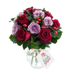 A luxurious, garden-inspired bouquet featuring an even blend of deep, velvety red roses and soft lavender roses, artfully packed into a round, abundant form that feels both opulent and intimate. Each rose is fully open, the layered petals catching soft daylight and revealing subtle variations - warm crimson, rich burgundy, pale lilac and dusty purple - that move the eye across the arrangement. Scattered among the blooms are small clusters of red-orange rose hips, their firm, glossy skins adding punctuated pops of colour and a rustic, seasonal texture. Lush, dark green leaves, likely Ruscus or Salal, weave through the roses to give structure and a verdant backdrop, lending cool contrast to the warm florals. The bouquet is displayed in a reflective mercury-style glass vase with a rounded base and a gently narrowed neck; its antiqued silver finish catches and diffuses highlights, while a length of natural twine is tied around the neck for an unfussy, handcrafted detail. A neat white gift tag dangles forward from the twine, marked with a small floral motif and soft script that suggests a personal greeting. Photographed on a pure white background, the arrangement reads as fresh and premium - a piece that conveys romance, thoughtful gifting, or quiet celebration and that would arrive beautifully presented for local delivery within Grove Park and neighbouring streets.