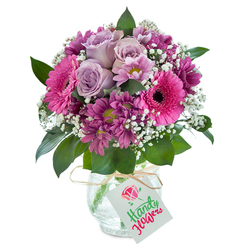 Bathed in gentle daylight, a lush bouquet of fresh, vividly coloured flowers sits nestled in a textured glass vase-a vision brought to life with the artistry of Grove Park's dedicated florists. The centerpiece features several flawless lavender roses, each unfurling gracefully to reveal a softness matched only by the welcoming spirit found near Grove Park station. Flanking the roses, dramatic Gerbera daisies in fuchsia pink boast open faces and rich, dark centers, their cheerful presence conjuring memories of strolls through Marvels Lane. Deep magenta chrysanthemums, some with a delicate hint of green at their hearts, fill out the edges, bringing depth and plushness to the design. Wispy white clouds of baby's breath hover throughout, bestowing an airy feel and echoing the light, relaxed ambiance of local parklands. Broad, glossy green foliage frames the entire arrangement, providing a natural contrast and grounding the palette in freshness. The clear glass vase features an eye-catching crackle effect, refracting light for a gentle, rustic touch that recalls the inviting windows of Grove Park cafés. At the neck, a simple twine of raffia supports a crisp white gift tag-an understated accent ready for a special message. This display is ideal for birthdays, celebrations, or simply to brighten a home, and is delivered with the trusted care and local knowledge for which Grove Park Florist is known.