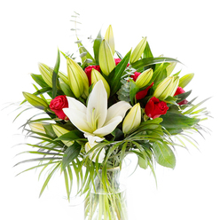 Set against a crisp white backdrop, this hand-composed bouquet centres on a single, fully opened creamy white Oriental lily whose satin petals unfurl with calm, sculptural grace, the pale yellow stamens and tender green pistil visible like a gentle heartbeat. Surrounding the focal lily are numerous lime-green and pale-yellow lily buds, some tightly furled and others just beginning to breathe open, creating a sense of anticipation and living movement in the vase. Scattered among the lilies are small, velvety red rosebuds-each a compact promise of passion-whose deep colour creates striking contrast and an intimate, romantic accent. The ensemble is embraced by diverse greenery: broad, glossy dark-green lily leaves that give solidity; long, feathery palm-like fronds in a fresher emerald that spill and arc to soften the silhouette; and rounded, silvery eucalyptus stems that introduce a cool, textured whisper between blooms. The arrangement sits in a clear, cylindrical glass vase where the pale green stems are visible through fresh water, a detail that reads as both honest and elegant under the studio lighting. The visual harmony-white purity, vibrant red drama, and layered greens-reads as tender and celebratory, ideal for anniversaries, birthdays, or to uplift a room. As a local touch, Grove Park Florist often prepares arrangements like this for delivery around Grove Park station and the Nature Reserve, promising fresh, thoughtfully staged bouquets that carry both scent and sentiment.
