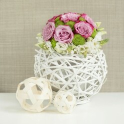 In this image, a finely balanced floral arrangement evokes the calm, sophisticated atmosphere of a Grove Park celebration, perhaps set just moments from the leafy quiet of Chiswick's riverside paths. The design centres on a spherical white vase crafted from slim, rattan-style twigs, woven into a delicate lattice that allows light to pass through, creating an interplay of shadow and brightness along the smooth white table surface below. Rising in a rounded, intentional dome from this airy base, an abundance of roses in blush pink and pale lavender tones draws the eye, each flower opened to show spiral layers of velvety petals, some edged with deeper pink accents that add a painterly touch. Between the roses, clusters of vivid green button chrysanthemums provide contrast and energy, their tight, bobbled heads and rich colour echoing the freshness of local Grove Park gardens in late spring. At the lower edge of the bouquet, soft white filler flowers emerge gently, forms small trails and clusters that slightly spill over the rim, giving the arrangement a relaxed, natural finish despite its carefully constructed shape. Resting on the same pristine white surface to the left of the vase are two decorative spheres of woven wicker, one large and one small, their pale, natural strands crossing and looping in an open, sculptural pattern that mirrors the vase's texture and reinforces the hand-crafted aesthetic. The background is a softly blurred expanse of warm beige fabric with gentle texture, a neutral canvas that allows the pink, green, and white tones of the flowers to stand out. The overall effect is of a beautifully composed wedding or dinner table centerpiece, suited to intimate gatherings in Grove Park where guests can appreciate both the floristry and the tactile, natural materials.
