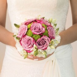 A moment of pure wedding-day anticipation is captured in this Grove Park scene: the bride, adorned in an off-white gown featuring intricate lace and gentle draping, holds a perfectly rounded bouquet that seems to glow with romance and celebration. Nestled at the heart are plush mauve roses, each petal unfurling with hints of fuchsia along the frayed edges, their colours reminiscent of a summer dusk over Grove Park's meandering paths. Surrounding the roses, crisp, lime-green pom-pom chrysanthemums add an unexpected, joyful burst-a fitting tribute to the garden feel found near the River Thames or up toward Staveley Road. The outer layer gleams with snow-white blooms, likely freesia or dainty spray roses, each cluster imbued with subtle fragrance and gentle grace. The bride's nails, finished in soft rosy polish and a glimmer on her ring finger, accent her hands as they cradle the bouquet, making every detail from florist's touch to personal adornment feel intentional. Soft, filtered light carries across the gown and petals, highlighting the interplay of colour and texture that defines expert floral design at Grove Park Florist. This image encapsulates the artistry and warmth found in hand-gathered wedding flowers, evoking the joy, care, and sense of promise at the heart of Grove Park celebrations.