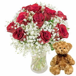 This image captures a warmly sentimental scene: a vase of deep red roses and a teddy bear arranged side by side against a bright white backdrop, the kind of thoughtful set you might see waiting for collection at Grove Park Florist. On the left, about twelve roses in a rich, velvety crimson shade stand tall in a simple, cylindrical glass vase. The petals, soft and layered, spiral outward from tight centres in various stages of bloom, some fully open and others still gently cupped, hinting at a bouquet that will continue to develop and change. Fine clusters of white baby's breath weave between the roses, their tiny blossoms forming a frothy halo that softens the strong red and adds a dreamy, romantic quality reminiscent of wedding flowers arriving in a quiet street off Grove Park Road. The stems are clearly visible through the water in the vase, their fresh green tones and occasional leaves adding texture and a sense of just-cut vibrancy. On the right, a medium-sized teddy bear in light brown plush sits upright with an easy, relaxed posture. Its fur appears soft and fluffy, inviting the idea of a comforting hug. Small dark eyes, an embroidered nose, and a subtle stitched smile give the bear a kind, approachable expression. A dark brown ribbon is tied neatly in a bow at its neck, complementing the deep tones of the roses and tying the composition together visually. The overall effect is one of classic romance and reassurance, the sort of gift you might send for an anniversary dinner in Grove Park, a birthday breakfast surprise, or a heartfelt apology delivered to a doorstep near local green spaces and tree-lined avenues.