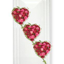 Three lush, heart-shaped arrangements dominate this serene composition, each one overflowing with roses in shades of deep red and luminous pink, creating a striking yet tender tribute. Arranged in a descending diagonal from the upper left to the lower right, the hearts appear almost like steps of affection, each linked to the next, as if charting the journey of love through life, loss, and remembrance. Every heart is packed tightly with blooms, their velvety petals layered so closely that the surface looks cushion-soft and rich with texture. Deep crimson roses provide the foundation, while clusters of brighter fuchsia, warm rose pink, and soft blush blooms weave among them, lending warmth and emotional nuance. The roses vary in openness, from tight buds to fully unfurled blossoms, which adds depth and a subtle feeling of movement, as though the tribute is still coming into full bloom. Around the edges of each heart, slender ivy vines curl and trail, their small, pointed leaves in a fresh, glossy green that outlines the hearts and extends downward to join them together. This delicate greenery brings a natural, almost woodland grace that feels at home in Grove Park, echoing the leafy paths and tree-lined roads near the station and the Thames. The hearts are set against a flawlessly white background, similar to a clean display board or wall in a local funeral venue, with soft, diffuse light falling evenly across the flowers, highlighting their colour and form without any harsh shadows. The mood is one of quiet devotion and respect, perfectly suited to a funeral service, graveside tribute, or memorial gathering for a loved one from the Grove Park community, where flowers are chosen with care to speak when words feel too small.