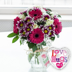On a pristine, light surface that could easily be a dining table in a Grove Park home, a meticulously arranged floral bouquet sits glowing in soft, natural light. The most striking elements are the gerbera daisies, broad-petalled and vivid in shades of fuchsia and magenta, their bold, dark centres providing a dramatic contrast and anchoring the entire design. Encircling these focal blooms are tight clusters of two-toned chrysanthemums, their petals transitioning from deep purple at the base to bright white at the tips, each blossom crowned with a golden-yellow centre that seems to sparkle. Creamy white roses, still modestly cupped with only a hint of their inner spirals visible, thread between the more vibrant flowers, adding a gentle, timeless elegance. Throughout the arrangement, clouds of tiny white gypsophila flowers drift like lace, softening the lines and heightening the bouquet's sense of fullness and abundance. Fresh green foliage, lush but not overpowering, frames the flowers and brings to mind leafy side streets and gardens near Grove Park station. The stems are clearly visible through the rounded glass vase, cut cleanly and standing in clear water, a small testament to the florist's care. Around the neck of the vase, a length of natural twine is tied into a neat bow, and from it hangs a white rectangular tag decorated with a stylised pink rose and cheerful green writing, suggesting a hand-crafted, made-with-care gift. Just to the right of the vase, a shiny, heart-shaped foil balloon catches the light, its silver border gleaming. The white centre is decorated with colourful, patterned letters in shades of pink, purple, and blue spelling out "Love n' Hugs 24-7," dotted with little hearts that amplify the affectionate theme. Against a bright, softly blurred background that hints at a sunny room, this pairing feels ideal for birthdays, anniversaries, or spontaneous gestures of love delivered across Grove Park.