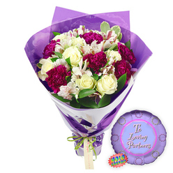 This image shows a jubilant graduation bouquet and balloon ensemble, posed against a bright white background as though ready to be carried out of a Grove Park florist and into a family celebration. At the heart of the bouquet, several creamy white roses stand proudly, their petals still slightly cupped and spiralled, capturing that moment between bud and full bloom that speaks of new beginnings. They are surrounded by clusters of deep fuchsia and magenta flowers, likely lush carnations or chrysanthemums, whose many-layered, ruffled petals create a vivid burst of colour and texture. Fine white filler flowers float between the larger blossoms, adding a light, airy quality, while lush green leaves weave through the arrangement, bringing an impression of freshness and life, much like the nearby greenery along the Thames Path. 

The flowers are wrapped in a bold purple paper, folded with care to form sharp angles and overlapping layers that cradle the bouquet. This wrapping gathers neatly at the base, where a decorative ribbon in green and white-possibly patterned or textured-encircles and secures it, creating a finished, celebratory look. The stems are hidden within the wrap, making the bouquet feel polished and easy to present. Just below and to the right of the bouquet lies a round purple Mylar balloon, its metallic surface reflecting soft highlights. In graceful white script across the centre, the message "To a Loving Graduate" is clearly visible, turning the arrangement into a personalised congratulation. Near the balloon, a small oval tag or sticker marked in green and white suggests branding or a quality seal from a local shop such as Grove Park Florist. Together, the bouquet and balloon capture the pride, joy, and hopeful energy of graduation day, making this a fitting image for gifts celebrating school leavers, university accomplishments, and exam success throughout Grove Park and the surrounding neighbourhoods.
