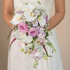 A modern Grove Park bride poses serenely, her arms enveloping an elaborate, cascading bridal bouquet-a floral work of art that captures the essence of riverside romance. Soft, natural light caresses the intricate lace of her gown, a canvas for the bouquet's myriad hues. Grand white Phalaenopsis orchids, with their silken petals and subtle yellow-gold centers, anchor the arrangement and exude quiet luxury. Tucked intimately among them are sculpted roses in delicate blush pink and pastel lavender, each bloom echoing the gentle tones of Chiswick's tranquil back gardens. Wisps of baby's breath, light as mist, create an airy fullness that gives the composition a sense of weightless beauty. From the bouquet's heart, shimmering fuchsia amaranthus trails downward, adding vivid drama and tactile intrigue, while clusters of small lavender blossoms and spring-green foliage weave through, suggesting freshness and possibility. The bride's poised hands cradle the bouquet centrally, forming a striking silhouette that feels both structured and freshly gathered-perfect for Grove Park weddings along Burlington Lane or at local stately homes. Every bloom and stem is balanced to evoke emotion, drawing on the artistry of Grove Park Florist's master design. This radiant bouquet, fresh and memorable, sets the stage for treasured wedding day memories throughout the neighborhood.