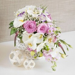A beautifully composed wedding-style arrangement spills from a round white wicker basket, as if set on a reception table in a Grove Park home overlooking the quiet streets around Burlington Lane. The flowers are densely arranged yet airy in feeling, led by large pastel pink roses that are open to their fullest, revealing spirals of silky petals with soft shadows catching in their folds. Between them glide white Phalaenopsis orchids, their broad, arching petals smooth and luminous, framing tiny yellow and pale orange centers that give the arrangement a delicate focal spark. Interspersed are white Lisianthus blooms, some edged with a soft lavender hue along their gently ruffled petals, bringing in an extra layer of romantic charm. Pale pink stock stems rise and tilt outward, giving the design gentle height and a hint of movement, as though the bouquet is quietly cascading toward the edge of the table. Fine clusters of white baby's breath are scattered throughout, filling small gaps with a lace-like, cloud-soft texture. Lush green foliage peeks out from between the blossoms, some leaves tucked deep inside, others spilling slightly over the basket's edge, suggesting fresh, garden-picked abundance. The white wicker basket itself sits on a smooth white surface that reflects a hint of light back into the flowers, while two decorative rattan or wicker spheres rest close by, echoing the basket's natural texture and adding a contemporary, handcrafted accent. A soft beige, linen-inspired background fades gently behind, creating a warm, neutral setting that allows the pinks, whites and greens to glow. The overall impression is of an elegant, thoughtfully crafted centerpiece, ideal for weddings, anniversaries or refined dinner parties throughout the Grove Park area.