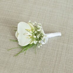 A refined white orchid boutonniere lies carefully arranged on a neutral, oatmeal-toned fabric, the weave of the cloth creating a subtle, homespun backdrop that recalls the relaxed charm of Grove Park's tree-lined streets. The focal point is a flawless Phalaenopsis orchid, its petals broad, smooth, and slightly glossy, curving outward like gentle wings. At the centre of the bloom, a whisper of soft yellow radiates from the throat, suggesting warmth and life, as if the flower has been freshly prepared in the cool of the morning at a local Grove Park workshop. Encircling this central orchid are airy tufts of baby's breath, each stem carrying dozens of tiny, snow-white flowers that cluster like a delicate mist. Their fragile, cloud-like presence softens the silhouette and brings a romantic, almost ethereal quality, ideal for a wedding at a nearby church or a civil ceremony in West London. Slim strands of vibrant green foliage thread through and beneath the flowers, their fine, fernlike fronds adding light movement and a breath of garden freshness reminiscent of walks near the Thames Path. The stems of the boutonniere are gathered with precision and bound in a crisp white satin ribbon, smooth to the eye and wrapped tightly enough to hold everything in perfect alignment. A small, neat bow marks the top of the binding, where a pin would slide through before resting on a suit jacket. The lighting in the image is soft and even, without harsh shadows, allowing the clean whites and fresh greens to stand out against the warm, tan fabric. This composition suggests a piece crafted for a groom, best man, or honored guest who appreciates classic, minimalist style - a discreet yet meaningful floral accent that pairs beautifully with tailored attire for weddings or formal events across Grove Park and the surrounding Chiswick area.