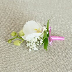 A single orchid boutonniere rests horizontally on a natural, oatmeal-toned linen fabric, the subtle weave of the cloth forming a calm, neutral stage for the flowers. At its centre, a large white Phalaenopsis orchid opens confidently, its broad petals satin-smooth and luminous, curving outward to reveal a delicately detailed throat tinged with soft yellow and the gentlest blush of pink. Around the orchid's base, tiny clusters of white baby's breath gather like a fine, airy mist, each miniature bloom adding lightness and texture, evoking the soft haze of an early morning stroll through Grove Park when the air still feels cool and clear. A few slender, fresh green leaves extend from beneath the blooms, their clean lines providing a crisp contrast that keeps the design feeling contemporary and uncluttered. Extending to one side, a short branching stem bears several tightly closed lime-green orchid buds, their smooth surfaces and rounded shapes suggesting life in pause, poised to open in days to come. The stems are carefully wrapped in a narrow band of bright fuchsia satin ribbon, tied into a neat, elegant bow whose glossy finish adds a hint of celebration and warmth. The vivid ribbon colour plays beautifully against the white petals and neutral background, giving the piece a subtle yet joyful accent, ideal for a groom, groomsman, or prom guest heading to a formal event in or around Grove Park. The soft, even lighting reveals every curve and texture, making this boutonniere feel intimate, thoughtful, and crafted with quiet precision and care.