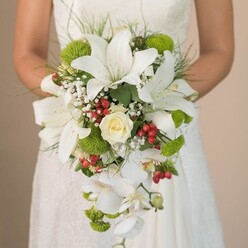 Bathed in golden daylight, this photograph reveals a Grove Park bride embracing a luxurious, teardrop-shaped cascading bouquet, its artistry speaking volumes about the skill of local florists. Striking white lilies-large and velvety, with just a dusting of reddish speckles-form an eye-catching focal point, their petals radiating grace and dignity. Nestled within, a creamy rose with perfectly curled petals adds a touch of soft tradition. Spiralling outward, whimsical lime-green cushion mums introduce a fresh air, their playful hues echoing the lively neighbourhood gardens nearby. Wispy sprays of pure white baby's breath weave among the main blossoms, diffusing the composition with lightness and a gentle cloudlike texture. Bright clusters of hypericum berries, painted a festive glossy red, dot the bouquet with cheerful contrast, reminiscent of the community's celebratory moments. The trailing edge of the bouquet is adorned with cascading white Phalaenopsis orchids, their silky petals arching in a waterfall of movement and sophistication. Elongated strokes of grass-like greenery and lush foliage frame the bouquet, offering a sense of wild elegance and reflecting the organic landscape surrounding Grove Park. The bride's hands cradle the stunning display against the textured lace of her gown, hinting at a story that unfolds between tradition and new beginnings. This elaborate arrangement, artfully designed to echo the spirit of events along Grove Park Terrace or near Chiswick House, brings a sense of timeless joy and local connection to any wedding day.