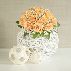 A sophisticated floral display is captured in soft, even daylight, showing a perfectly rounded cluster of peach and apricot roses poised above an airy white lattice vase. Around twenty to thirty roses are massed together, their petals arranged in a gentle dome that feels both abundant and controlled. Each rose carries a warm gradient of colour: a richer, sunset-orange heart that softens outward into delicate, almost pastel-peach tips. Some blooms are just beginning to unfurl, with spiralled centers still tight and promising, while others are more open, revealing the full, velvety whorl of petals that a seasoned florist in Grove Park might choose for a special celebration. Fine sprays of white gypsophila weave between the roses, their tiny blooms creating a mist-like border that lightens the design and adds a sense of breath and movement. Dark, glossy green leaves tuck around the base of the bouquet, grounding the pastel tones and lending a natural structure. The flowers emerge from a striking spherical container crafted from slender white strands, intertwined and knotted into an open, geometric globe that feels modern yet handmade, like a carefully curated piece from a local Chiswick or Grove Park design studio. On the white table in front of the vase sit two matching decorative orbs in the same string-like material, one larger and slightly patterned in neat segments, the other looser and more free-form, echoing the main piece without competing with it. The background is a pale beige or greige wall, quietly textured, offering just enough visual interest without drawing attention away from the centerpiece. Altogether, the composition evokes a calm, contemporary atmosphere, ideal for an elegant dinner party, wedding reception, or home staging for a refined property near Grove Park station, conveying care, craftsmanship, and a gentle sense of celebration.