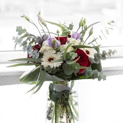 Presented in a transparent glass vase atop a sun-drenched table near Grove Park's leafy avenues, this bouquet expresses quiet confidence and local flair. At first glance, bold red roses capture the eye, their velvety richness harmoniously softened by luminous white gerbera daisies with deep espresso centers. Eucalyptus stretches outward in graceful arcs, its silvery leaves shimmering with the green of spring mornings by the Thames Path. Soft mauve lisianthus blooms, many still partly closed, thread gentle promise throughout-a fitting tribute for celebrations, remembrance, or gestures of comfort within the Grove Park community. Sprays of small, white filler blooms add plush fullness, clustering like morning clouds over the arrangement. Embracing it all are structured, broad green leaves, lending a finishing polish and frame. Through the vase's glass, vibrant stems are visible, resting in water, a pleasing detail for those who value both health and visual delight in cut flowers. Subtle scents of rose and eucalyptus intermingle, creating a backdrop both refreshing and meditative. Whether the bouquet is destined for a birthday table along Grove Park Terrace or as a thank you gesture in a neighbor's home, its balanced composition reflects the enduring ties and artistry cherished by Grove Park Florist.
