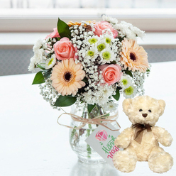 On a bright white tabletop that catches a faint reflection, a delicate anniversary arrangement is thoughtfully styled, reminiscent of a lovingly prepared gift awaiting its recipient in a Grove Park townhouse near Chiswick Main Line station. In the centre, a clear glass jar vase displays an artfully balanced bouquet of pastel flowers. Soft pink roses with smooth, layered petals rest beside peach gerbera daisies, whose dark chocolate brown centres anchor the composition. Scattered between these larger blooms are filmy sprigs of white baby's breath and tidy clusters of white chrysanthemums with lime-green hearts, creating both fullness and a sense of lightness. Around the rim of the jar, a length of natural twine is wrapped and tied into a modest bow, from which a small white tag is suspended. On the tag, a pink heart icon and flowing green script reading Handmade speak to the personal touch and attention given to each detail by a careful florist. To the right, perched on the same immaculate surface, a cream teddy bear sits upright, as if keeping affectionate watch over the flowers. The bear's fur looks plush and slightly shaggy to the touch, with dark, bead-like eyes, a soft brown nose, and a fine brown satin ribbon tied neatly at its neck, adding a subtle hint of formality. The backdrop is softly out of focus, suggesting daylight pouring in from a nearby window and lending the tableau a calm, intimate atmosphere. This pairing of gentle flowers and a cuddly bear radiates warmth, making it perfectly suited to marking an anniversary in Grove Park with a quiet yet meaningful gesture of love.