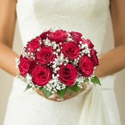 Amid Grove Park's leafy tranquility, a bride stands poised and serene-her hands cradling a bouquet overflowing with velvety deep red roses, each bloom crafted to perfection in homage to enduring romance. The flowers, gathered into a domed arrangement, are interspersed with frothy white gypsophila, whose petite clusters provide a graceful counterbalance to the saturated richness of the roses. Lush green leaves at the bouquet's base impart a fresh, natural accent, reminiscent of the thriving gardens along Grove Park Road. The bride's polished nails and gentle grip express an intimate anticipation, while the intricately textured wedding dress-suggesting lace or subtle embroidery-adds further elegance. Cascading to the right, a soft sash or fabric panel hints at the movement and joy of a day unfolding. The background is artfully diffused into creamy light and beige, allowing the bouquet's hues and intricate structure to shine without distraction. This arrangement, designed by Grove Park Florist for weddings in the area or delivery to locations near Chiswick and Bromley, embodies classical sophistication with a local twist-ideal for those envisioning a wedding steeped in romance and timeless style, guided by expert hands familiar with Grove Park's community traditions.