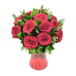 This detailed studio image shows a classic, deeply romantic bouquet of approximately a dozen to fifteen red roses displayed in a rich red glass vase, set sharply against a pristine white background that lets the colours sing. Each rose is at the perfect stage of bloom: petals fully unfurled into tight, intricate spirals, yet still plump and velvety, suggesting lasting freshness and care in selection. The red hue is intense, closer to garnet or ruby than scarlet, creating a sense of luxury and heartfelt emotion. Around and between the rose heads, a generous layer of dark green foliage, most likely ruscus or similar greenery, introduces contrast and structure; the leaves are smooth, glossy, and slightly pointed, catching little highlights of light that lend the arrangement depth and dimension. The flowers are composed into a rounded, almost dome-shaped design, a style favoured by many Grove Park couples for anniversaries and romantic gestures, with each bloom facing outward so the viewer can appreciate every perfect petal. The red glass vase has a gently flared rim and curves inwards as it descends, tapering to a narrower base. Its translucent, ribbed or softly swirled surface reflects the surrounding light, producing subtle variations in red that echo the richness of the petals above. The stems inside are only faintly visible, adding to the sense of cohesion between vase and flowers. Bright, even illumination from the front and sides casts soft shadows beneath the vase, grounding it while keeping the focus firmly on the roses. The composition conveys quiet intensity and sincerity, like a bouquet prepared for a meaningful evening at home in Grove Park, perhaps after a walk through the nearby green spaces, when words alone don't feel quite enough.