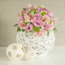 On a clean white surface that could easily be a Grove Park kitchen island or hallway console, a generous floral centerpiece radiates soft colour and calm, framed by a subtly textured light beige wall that keeps all focus on the flowers. At the heart of the scene is a striking white vase, rounded and slightly bulbous, formed from white strips interlaced into an open, lattice-style pattern, giving it a handcrafted, contemporary-rustic character. It feels solid yet airy, like a sculptural basket reimagined for modern interiors around Chiswick or Lee. Overflowing from this vessel is a carefully balanced bouquet of pastel pink and lavender roses, their petals velvety and lush, with some blooms fully open and others just beginning to unfurl, suggesting movement and life. Between them, elegant pink alstroemeria lift their slim, trumpet-shaped flowers, each petal brushed with fine streaks of deeper pink or soft golden tones at the throat, lending the bouquet a painterly detail best appreciated up close. Punctuating the soft pastels are bright lime green button chrysanthemums, compact and spherical, adding playful contrast and energy while drawing the eye across the arrangement. Delicate clusters of white baby's breath weave through everything, like tiny clouds suspended between stems, creating a gentle halo around the main blooms. Deep green foliage is tucked throughout, adding structure, freshness, and a hint of woodland fragrance. To the left of the vase rest two decorative spheres fashioned from natural-toned, woven strips, echoing the pattern of the vase and adding balance and rhythm to the composition. The overall impression is of quiet celebration and refined taste: a piece that could brighten a Grove Park living room for an anniversary, a new home, or a softly spoken "thank you," arranged with the considered touch of a local florist who understands how flowers can change the mood of a room.