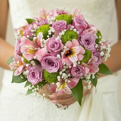 This Grove Park-inspired bridal bouquet brims with elegance and sentiment, delicately held in a bride's hands and set against a whisper-soft veil of white lace. Blushing lavender and pink roses, symbols of everlasting love, form the bouquet's core, each bloom unfolding gently like petals newly opened along the gardens bordering Grove Park. Interspersed among them, vivid lime green button chrysanthemums add a fresh, modern twist, their distinctive pom-pom shapes sparking visual interest reminiscent of early spring on Fauconberg Road. Elegant pink alstroemeria lilies introduce subtle complexity, their trumpet-shaped petals brushed with maroon and dappled with yellow, evoking both the familiar and the exotic. Wisps of baby's breath weave a misty layer throughout, creating a sense of airiness and tranquil joy that echoes the leafy calm of local parks. The bouquet's dark green foliage amplifies its natural beauty, providing structure and harmony, while every detail is illuminated by gentle, natural light. Seen in close detail, the bouquet's texture and colour blend is a testament to its careful creation, the result of Grove Park Florist's dedication and creativity. Perfect for weddings set against the borough's historic backdrops, this floral design evokes hope, romance, and the distinctive charm of Grove Park's communities, marking a new chapter with grace and joy.