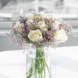 A round, luxuriously full bouquet of fresh white roses and lavender statice is captured in soft daylight, arranged with the kind of quiet precision a local Grove Park florist would bring to a special order. At the centre, roughly a dozen creamy white roses form a generous cluster, their petals unfolding layer by layer in perfect spirals. The tone is not stark white but a gentle cream that hints at ivory, giving each bloom a warm, flattering glow rather than a harsh brightness. Cradling these roses is a halo of lilac-tinted statice, made up of countless tiny blossoms that resemble a fine, cloud-like lace. Their muted lavender colour sits delicately between grey and purple, adding depth and a whisper of colour without disturbing the bouquet's calm, neutral palette. The mix of textures is striking: the roses appear plush and almost velvety, while the statice feels dry and papery, creating a subtle interplay of softness and airiness. All the stems are gathered in a tall, cylindrical clear glass vase, where straight green stems and their natural curves can be clearly seen beneath the waterline. The water itself looks crystal clear, catching the light along the rim of the vase. Set on a clean white surface that gently reflects the arrangement, the bouquet is bathed in diffused daylight, likely from a nearby window overlooking a quiet Grove Park side street or the greenery near Grove Park station. The blurred, pale background enhances the sense of serenity, making this arrangement perfectly suited to an intimate anniversary breakfast, a minimalist wedding setting, or a simple gesture of appreciation delivered to a loved one's doorstep in the local area.