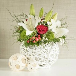 A beautifully composed centerpiece stands ready to anchor a wedding table in Grove Park, its carefully chosen flowers arranged in a distinctive round white vase with an open, basket-like weave. This sculptural container cradles a generous grouping of fresh white lilies, their tall stems and elongated buds reaching upwards while several blossoms have already opened wide, petals curling back elegantly to expose delicate markings and golden stamens at their heart. Interlaced with the lilies are sumptuous red roses, their velvety petals layered tightly in classic spirals; one central rose in particular draws the gaze, a rich crimson focal point that feels made for a romantic toast or anniversary dinner near Chiswick's riverside. Clusters of bright green, spherical flowers-likely green dianthus or chrysanthemums-sit like soft cushions among the blooms, adding an unexpected yet harmonious texture and a vivid, mossy hue. Fine sprays of gypsophila thread lightly throughout the arrangement, their tiny white blossoms forming a gentle mist that bridges the bold colours. Small clusters of glossy red berries peek from between the foliage, lending a subtle seasonal touch that would suit winter weddings or holiday tablescapes around Grove Park. The foliage itself ranges from feathery, wispy fronds that arc loosely outwards to broader, deeper green leaves that anchor the design, giving the bouquet structure, depth, and a sense of natural abundance. To the left side of the vase rest two decorative woven balls in a soft natural tone, their open lattice echoing the vase and completing the scene with contemporary flair. Behind it all, a softly textured neutral wall in light beige and grey ensures the red, white, and green tones stand crisp and clear, creating a refined, inviting mood for gatherings with family and friends.