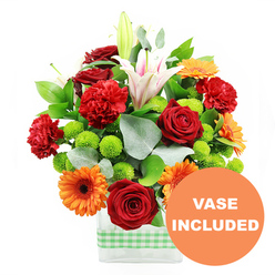 This photograph presents a joyful, garden-inspired bouquet displayed in a modern, clear rectangular vase, the base wrapped with a neat band of light green and white gingham ribbon that evokes a relaxed, kitchen-window feel familiar in Grove Park homes. Above the glass, the flowers form a dense, rounded arrangement where warm reds and oranges are balanced by crisp whites and lively greens. Deep red roses, their petals velvety and tightly packed, serve as romantic focal points. Nestled alongside, red-orange carnations with soft, ruffled heads lend a plush, comforting texture. Bright orange gerbera daisies punctuate the design, each with a dark central eye and a halo of flat petals that draw the gaze outward. Emerging gracefully through the middle, white Oriental lilies add vertical interest; some are fully open, revealing pale pink veins and prominent stamens, while others are still in elongated buds, promising continued flowering. Interwoven throughout are clusters of lime-green button chrysanthemums, their tiny spherical blooms offering a playful contrast in both colour and form. The arrangement is richly backed by glossy deep green leaves and airy sprigs of eucalyptus in cool silver-green tones, which introduce gentle curves and movement. The stems can be seen clearly through the vase, immersed in water that catches the light and hints at freshness. In the bottom right corner, a bright circular orange graphic slightly overlaps the vase and carries the message VASE INCLUDED in bold white lettering, making it clear that this is a ready-to-display gift. The overall impression is of an energetic, uplifting bouquet that would brighten a dining table near Chiswick Station, a studio overlooking the Thames, or arrive as a same-day delivery from Grove Park Florist to mark joyful news or heartfelt appreciation.