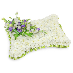 A carefully composed floral pillow, shaped like a gentle rectangular cushion, is crafted from a dense field of immaculate white chrysanthemum heads so closely arranged they create a seamless, velvety surface. The overall effect is one of serene softness: each little chrysanthemum contributes to a unified white plane that suggests rest, purity, and solace. The pillow's softly rounded corners and slightly elevated centre give it a restful, lifelike silhouette, and a soft shadow beneath anchors the piece against a clear white backdrop.

Framing the piece is a neat pleated trim of light green ribbon, folded into tender scallops that trace the pillow's perimeter and lend a fresh, hopeful edge to the composition. In the top-left corner a small, artful spray of accent flowers provides quiet contrast - pale white lisianthus bloom beside rich purple lisianthus, with a few buds tucked in for depth. Interspersed fern fronds and glossy dark leaves add texture and movement, while a broader green leaf wrap at the base holds the cluster with understated grace. The combined scent is muted and clean, a soft mix of chrysanthemum freshness and the faint sweetness of lisianthus.

Designed for moments of remembrance, this sympathy cushion is presented as a dignified tribute suitable for funerals or graveside placement. Made with care for families in Grove Park and nearby communities such as Sundridge Park and Mottingham, it conveys quiet respect and comforting presence when honouring a loved one.