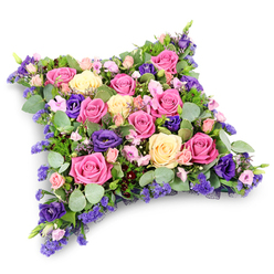 This image captures a beautifully composed cushion-shaped funeral tribute, designed to look like a soft pillow laid out in quiet reverence. The arrangement forms a neat rectangular cushion with gently rounded corners, shimmering against a clean white background that enhances its rich colours and textures. The pillow's surface is almost entirely made up of roses in shades of fuchsia, blush pink, and creamy yellow, their petals overlapping in tight formation to create a plush, padded effect. Some roses are fully open, displaying their layered hearts, while others are still partly furled, hinting at the natural unfolding of time and memory. Between them, deep royal purple lisianthus blooms emerge, their velvety petals adding contrast and an air of solemn elegance, reminiscent of the calm, tree-lined paths around Grove Park. Tiny lavender-lilac filler flowers, possibly statice or limonium, weave across the tribute in gentle clouds of colour, softening the transitions between the larger faces of the roses. Nestled beneath and around the flowers, rounded, silvery eucalyptus leaves catch the light with a muted sheen, while darker, feathery green foliage adds depth and neatly frames the edges, ensuring the cushion's outline remains well-defined. The overall mood is tranquil and reflective, ideal for expressing condolences at a funeral, burial, or cremation service in Grove Park or the surrounding neighbourhoods. The tribute feels freshly prepared, as though crafted just moments ago by an experienced local florist, offering families a graceful way to say goodbye when it matters most.