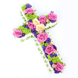 The photograph shows a striking floral cross laid out on a luminous white background, its form captured from above with a slight diagonal tilt that adds energy and depth to the composition. A border of small white spray chrysanthemums tightly encircles the cross, creating a soft, cloud-like frame that feels both pure and comforting. Beneath this plush edge, a light green ribbon or folded foliage peeks through, tracing the perimeter with a subtle band of fresh colour reminiscent of the trees around Grove Park and the nearby Thames path. Inside this frame, the cross is richly filled with flowers in a harmonious, yet vivid palette. Large, fully opened roses in hot pink and gentle buttery cream are spaced along the arms and length of the cross, drawing the eye from top to base. Their layered, velvety petals give the tribute a luxurious, heartfelt character. Between the roses, bright lime-green button chrysanthemums cluster in rounded groups, their compact, textured forms adding a cheerful contrast that brings modern flair to this traditional symbol. Deeper in the design, petite purple and violet blooms-likely lisianthus or statice-thread through the spaces, lending depth and a quiet, reflective tone suitable for a funeral or memorial service. The daylight-style lighting is clear and even, allowing each petal, button flower, and ribbon fold to be seen with clarity. Crafted with the care of an experienced local florist, this floral cross feels at home in Grove Park churches and chapels, an uplifting Christian tribute that combines faith, remembrance, and a gentle celebration of life.