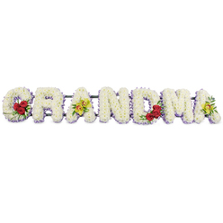 A lovingly crafted sympathy arrangement spells out the word GRANDMA in large floral letters, captured against a plain, bright white background that makes the colours and textures stand out clearly. Each letter is generously shaped with dense clusters of crisp white chrysanthemums, their small, rounded petals packed so closely that the surface appears soft and uniform, like a carefully smoothed blanket of blooms. Just under the white flowers, along the base of every letter, runs a narrow band of pale lavender or light mauve blossoms, which provides a gentle outline and hints at depth without disturbing the overall serenity of the design. On the opening G, a vivid cluster of three deep red roses sits on the lower left curve, framed by rich, dark green leaves that bring structure and contrast. The first A is dressed with two yellow-green cymbidium orchids laid across its bar, their fresh chartreuse tones and speckled centres giving an elegant, modern touch. The letters R, N, and D remain purely white with the soft lavender edging, offering a tranquil rhythm through the centre of the tribute. The M then introduces another grouping of three red roses in its lower right arch, a visual echo of the G, and the final A mirrors the first by displaying two more chartreuse orchids, perfectly balancing the composition. A discreet, dark metal frame connects the letters along the top, sturdy yet barely visible. The overall effect is one of calm respect and heartfelt love, the kind of personalised floral tribute that might be placed at a funeral service or graveside in and around Grove Park, offering family and friends a gentle, beautiful focus for their memories of a cherished grandmother.